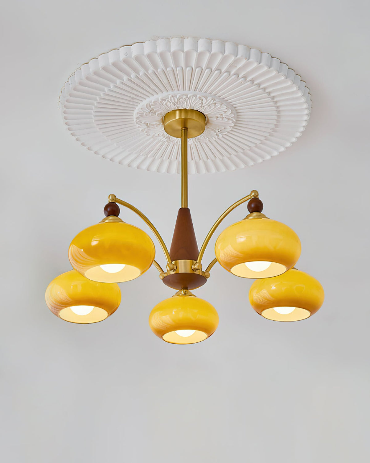Five lit caramel glass shades, with gold caps and wooden finials, on this Clérachic chandelier beneath a pleated white ceiling rose.