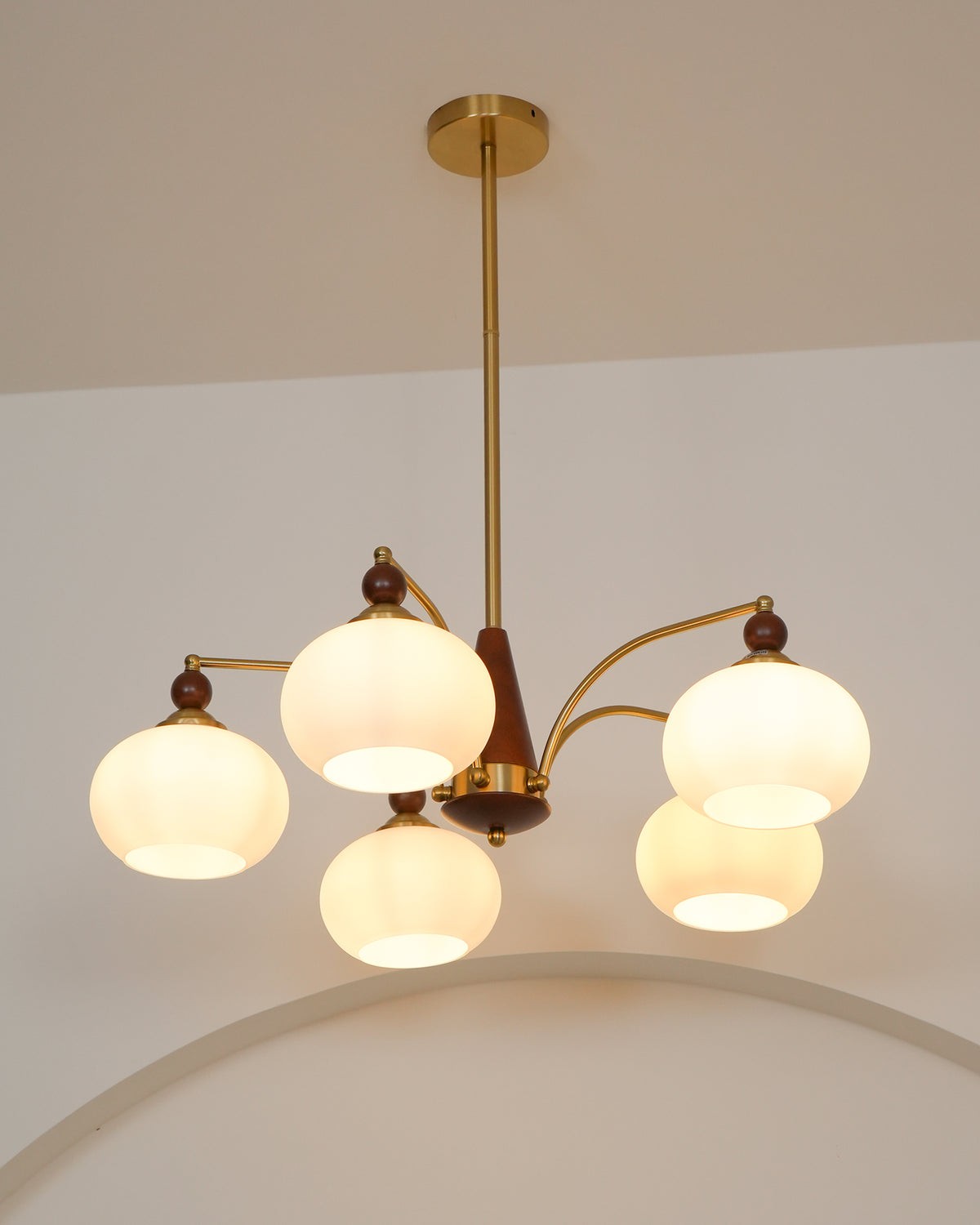 Lit white glass shades on this Clérachic chandelier, hanging from a gold rod beside a curved arch in the wall.
