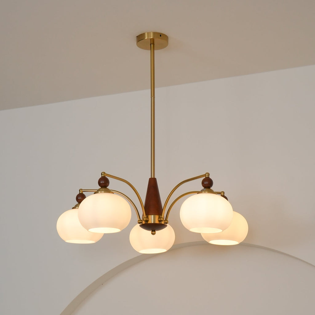 Front view of a Clérachic chandelier with five white glass shades lit warm, on a gold rod, against a pale wall.