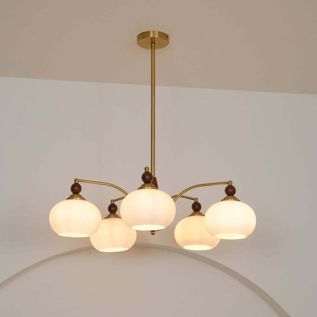 A Clérachic chandelier with five lit white glass shades seen from slightly below, with a gold ceiling plate above.