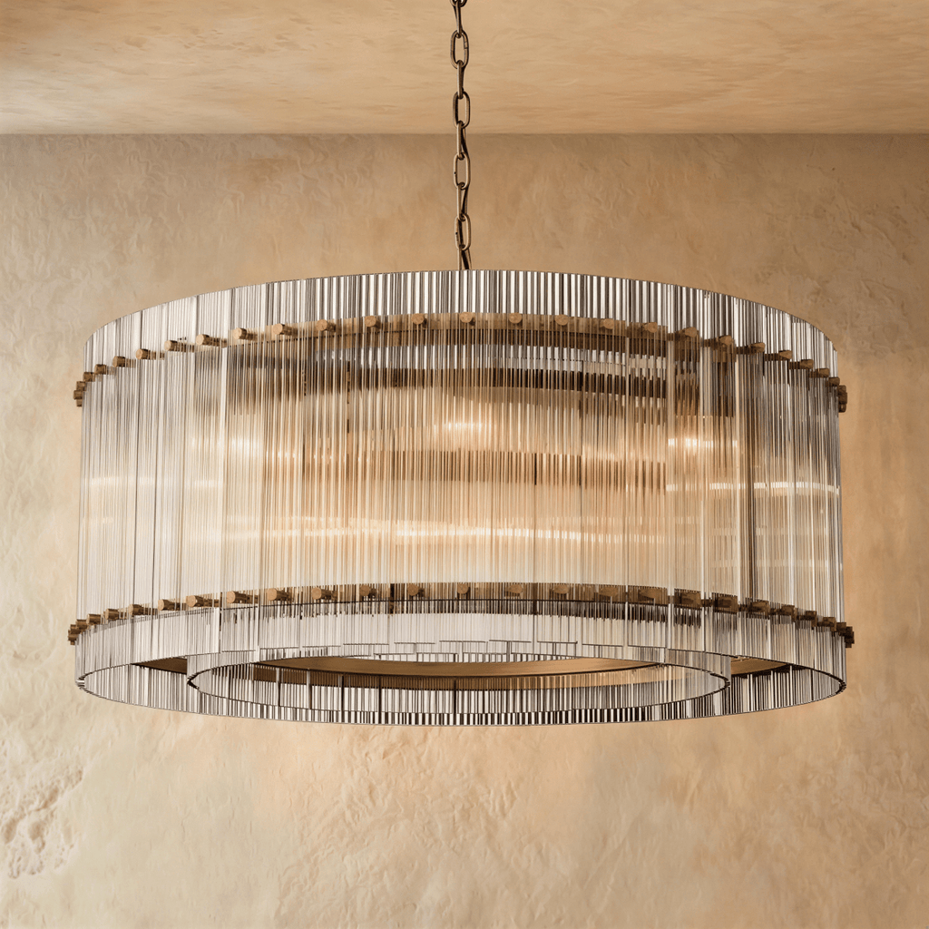 Clérachic wide drum chandelier of clear fluted glass rods with antiqued brass studs, hung on a chain against warm plaster.