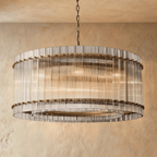 Clérachic wide drum chandelier of clear fluted glass rods with antiqued brass studs, hung on a chain against warm plaster.