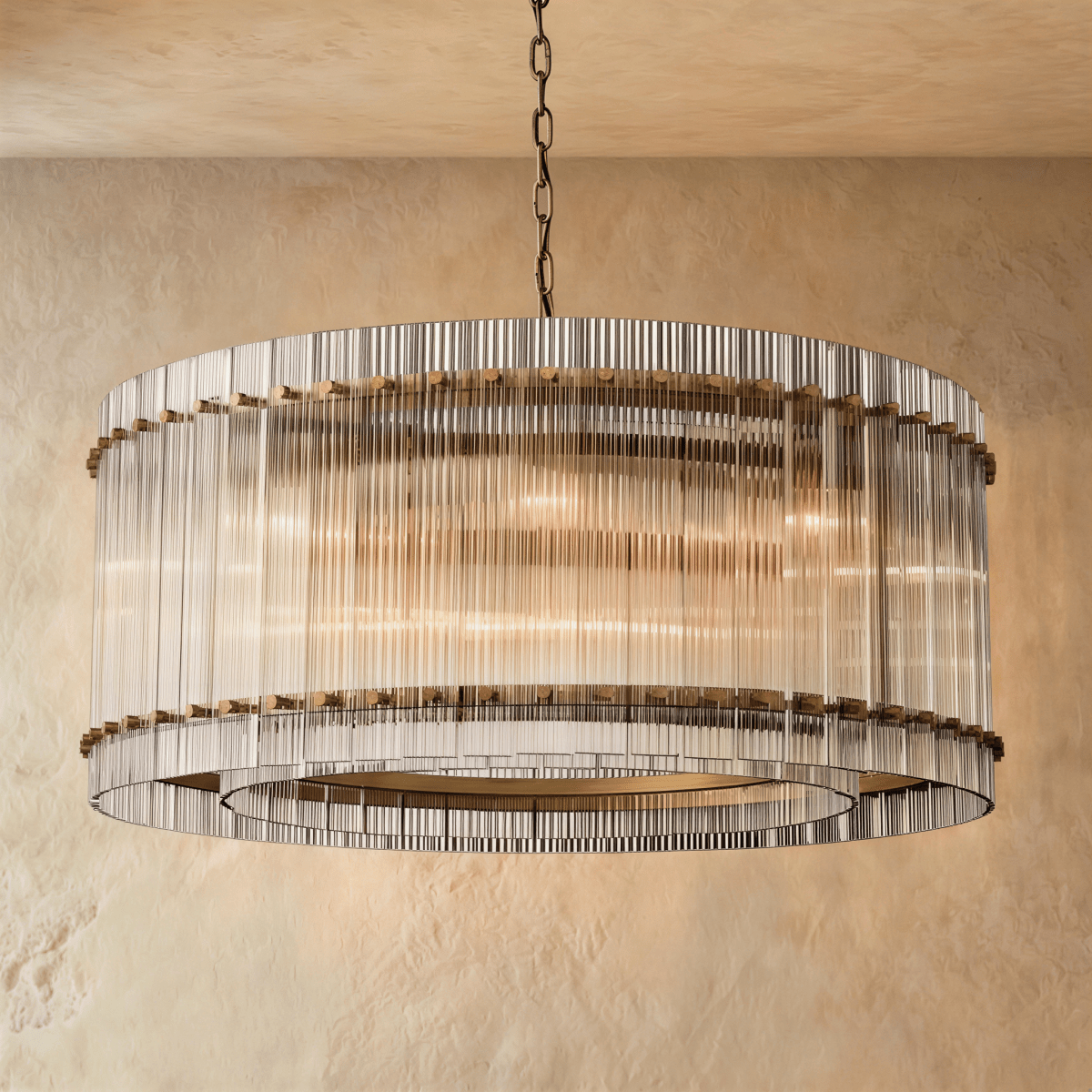 Clérachic wide drum chandelier of clear fluted glass rods with antiqued brass studs, hung on a chain against warm plaster.