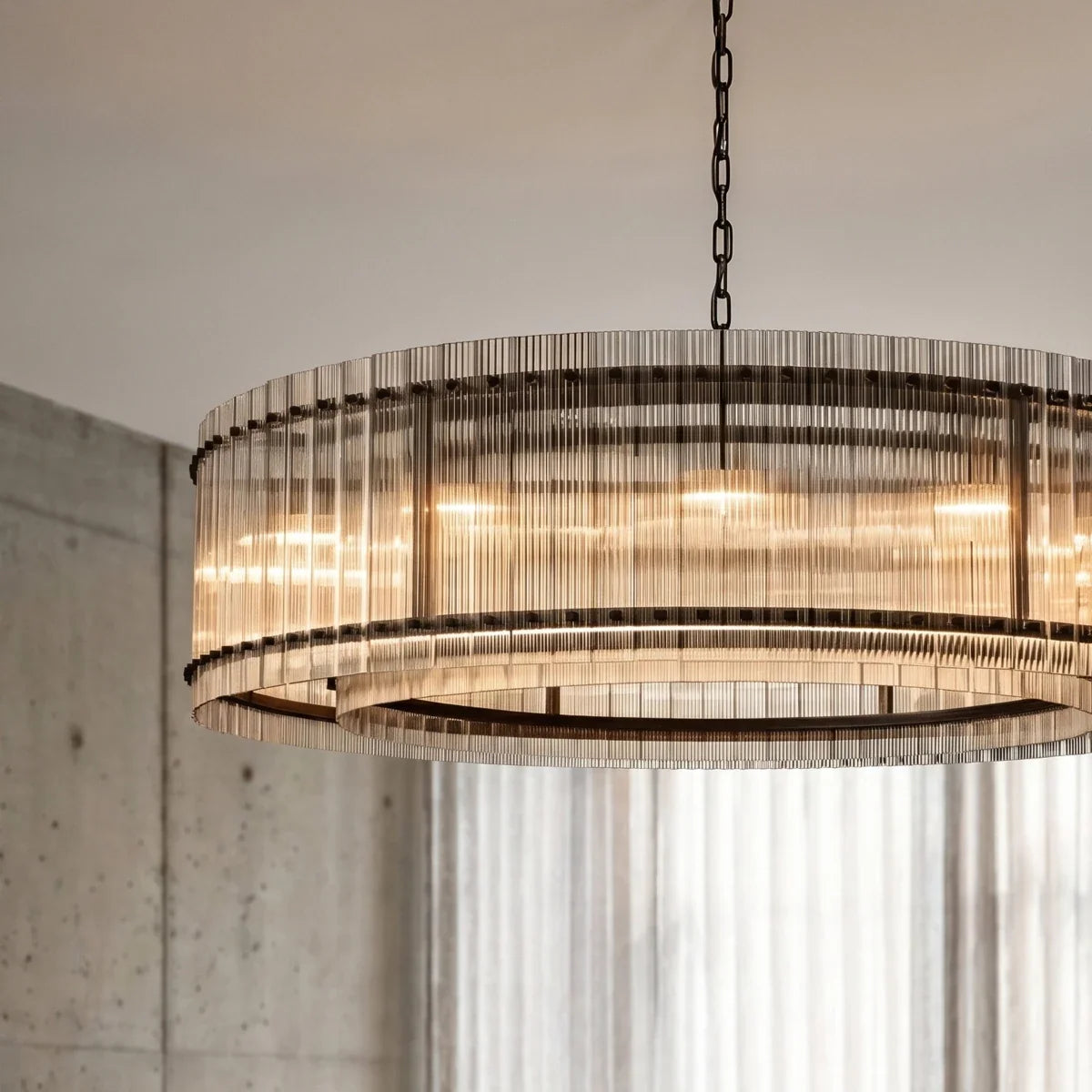 Clérachic fluted glass drum chandelier with dark metal studs and a glowing inner ring, seen close up against a concrete wall.