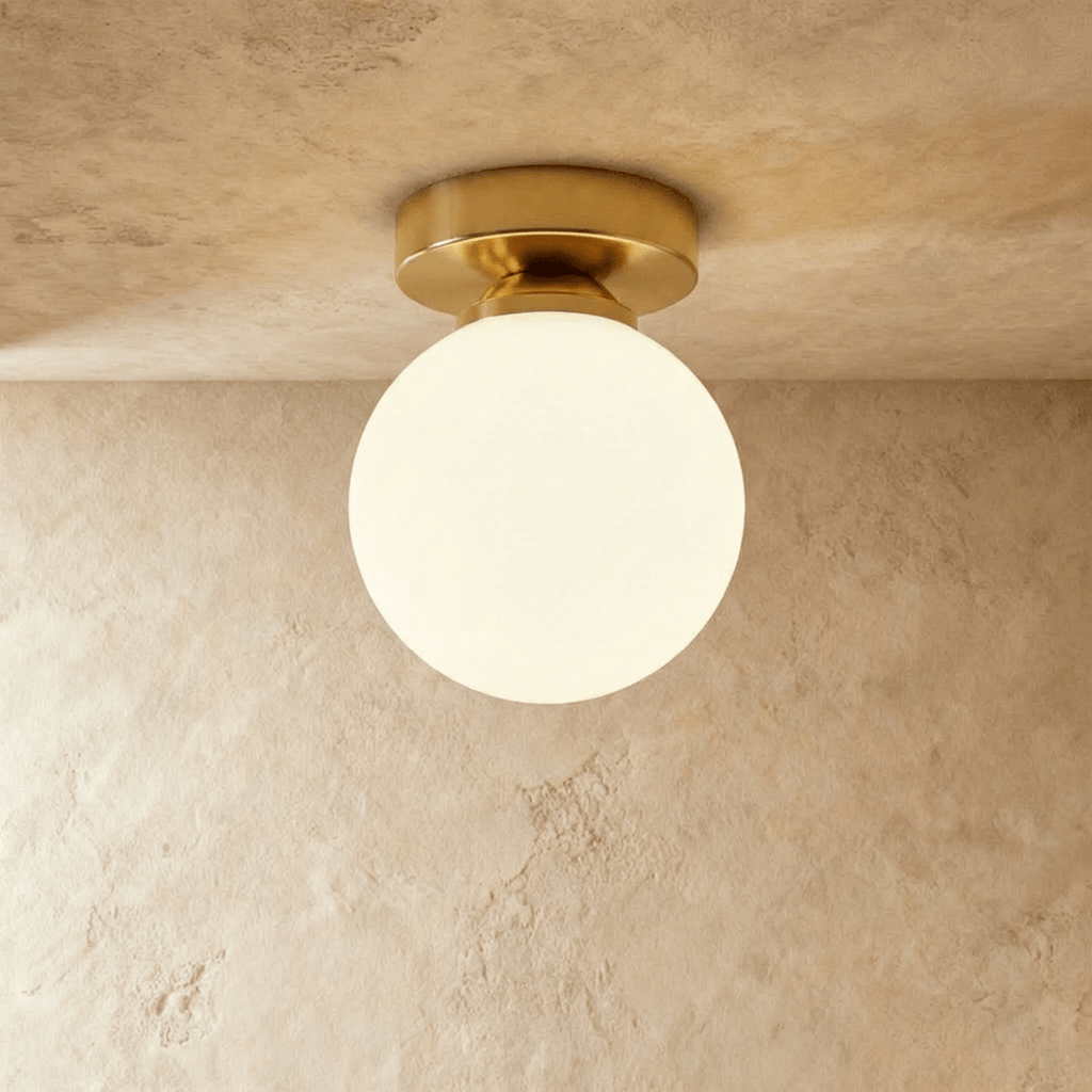 Clérachic flush-mount light in brushed gold, a single opal glass globe glowing warm on a plaster ceiling.