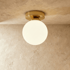 Clérachic flush-mount light in brushed gold, a single opal glass globe glowing warm on a plaster ceiling.