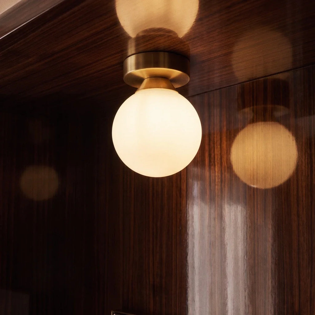 Clérachic flush-mount light detail in brushed gold, the opal glass globe glowing against wood panelling.