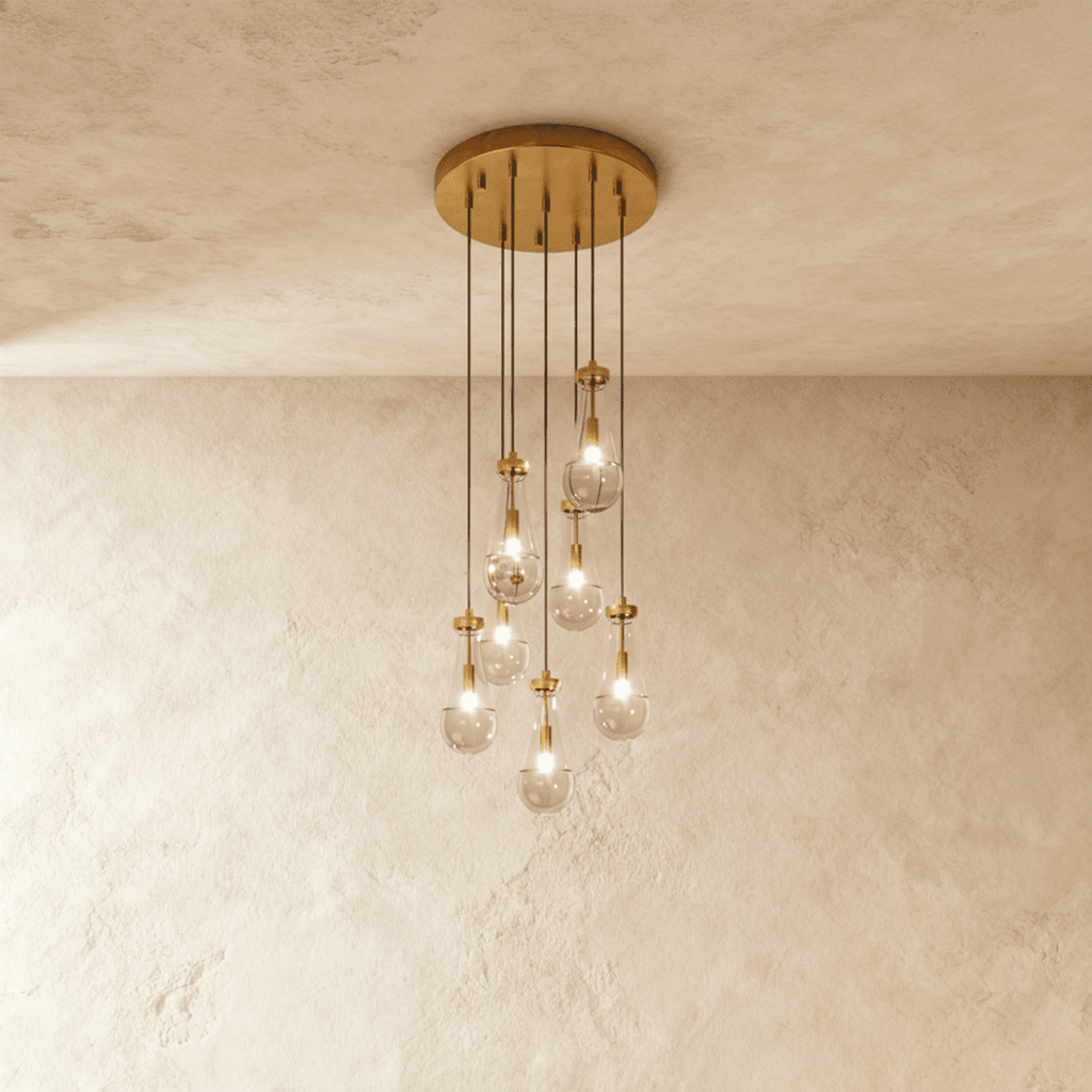Clérachic chandelier with 7 clear glass teardrops on thin cords from a round brushed gold canopy, glowing warm, against limewashed plaster