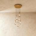 Clérachic chandelier with 7 clear glass teardrops on thin cords from a round brushed gold canopy, glowing warm, against limewashed plaster
