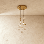 Clérachic chandelier with 7 clear glass teardrops on thin cords from a round brushed gold canopy, glowing warm, against limewashed plaster