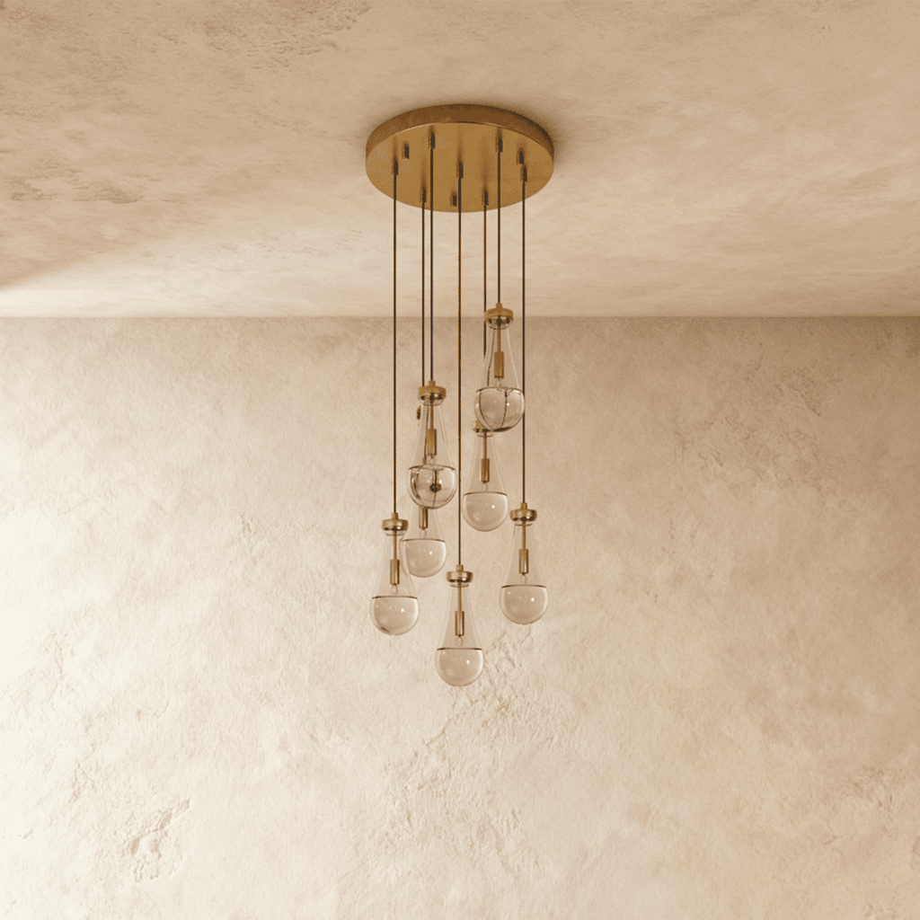 Clérachic chandelier with 7 clear glass teardrops on thin cords from a round brushed gold canopy, switched off, against limewashed plaster