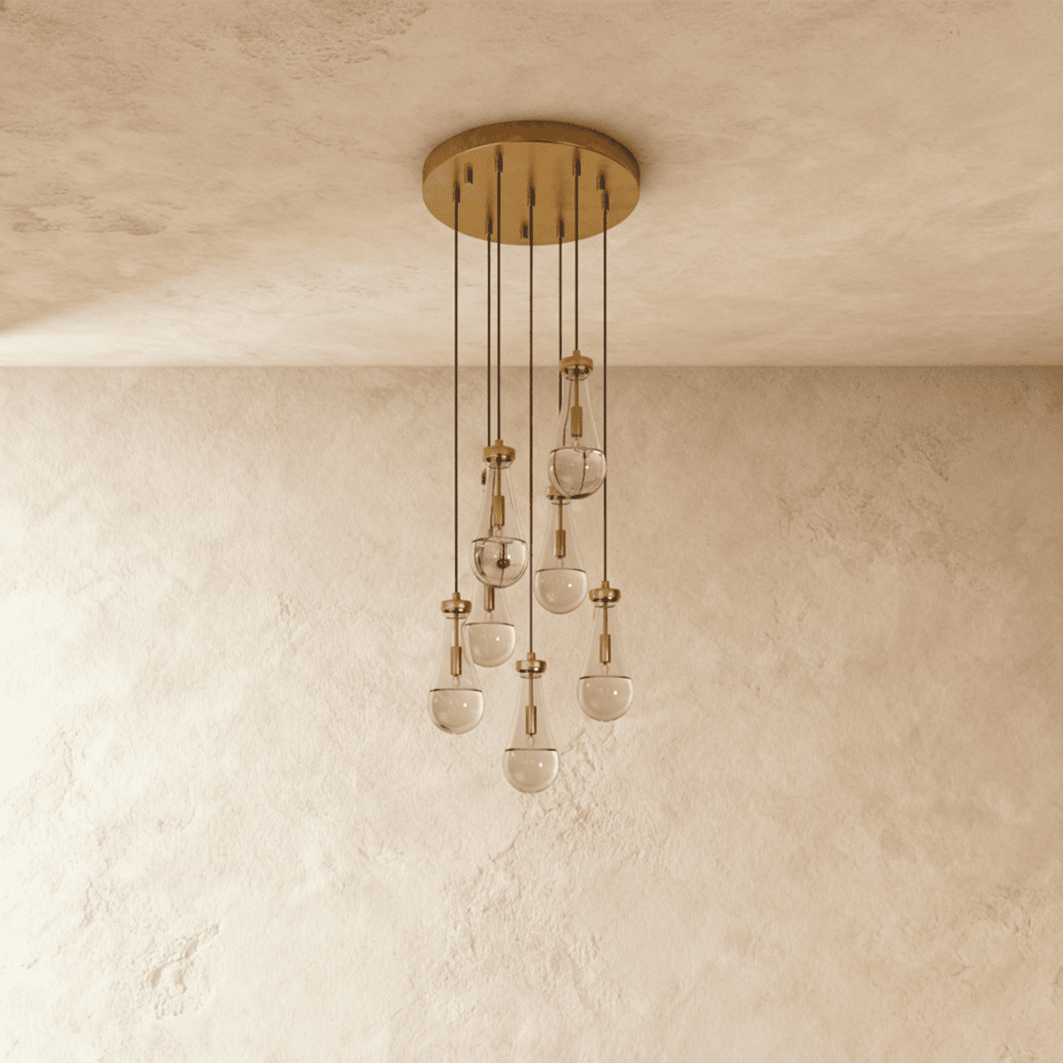 Clérachic chandelier with 7 clear glass teardrops on thin cords from a round brushed gold canopy, switched off, against limewashed plaster