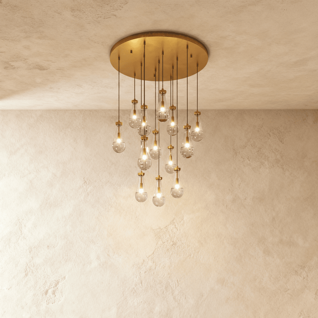Clérachic chandelier with 13 clear glass teardrops on thin cords from a round brushed gold canopy, glowing warm, against limewashed plaster