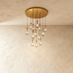 Clérachic chandelier with 13 clear glass teardrops on thin cords from a round brushed gold canopy, glowing warm, against limewashed plaster