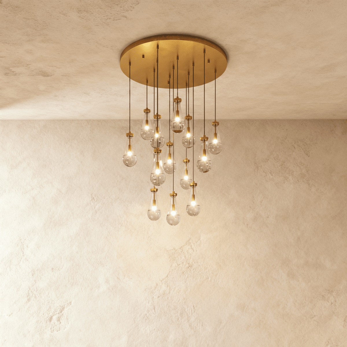 Clérachic chandelier with 13 clear glass teardrops on thin cords from a round brushed gold canopy, glowing warm, against limewashed plaster