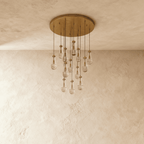Clérachic chandelier with 13 clear glass teardrops on thin cords from a round brushed gold canopy, switched off, against limewashed plaster