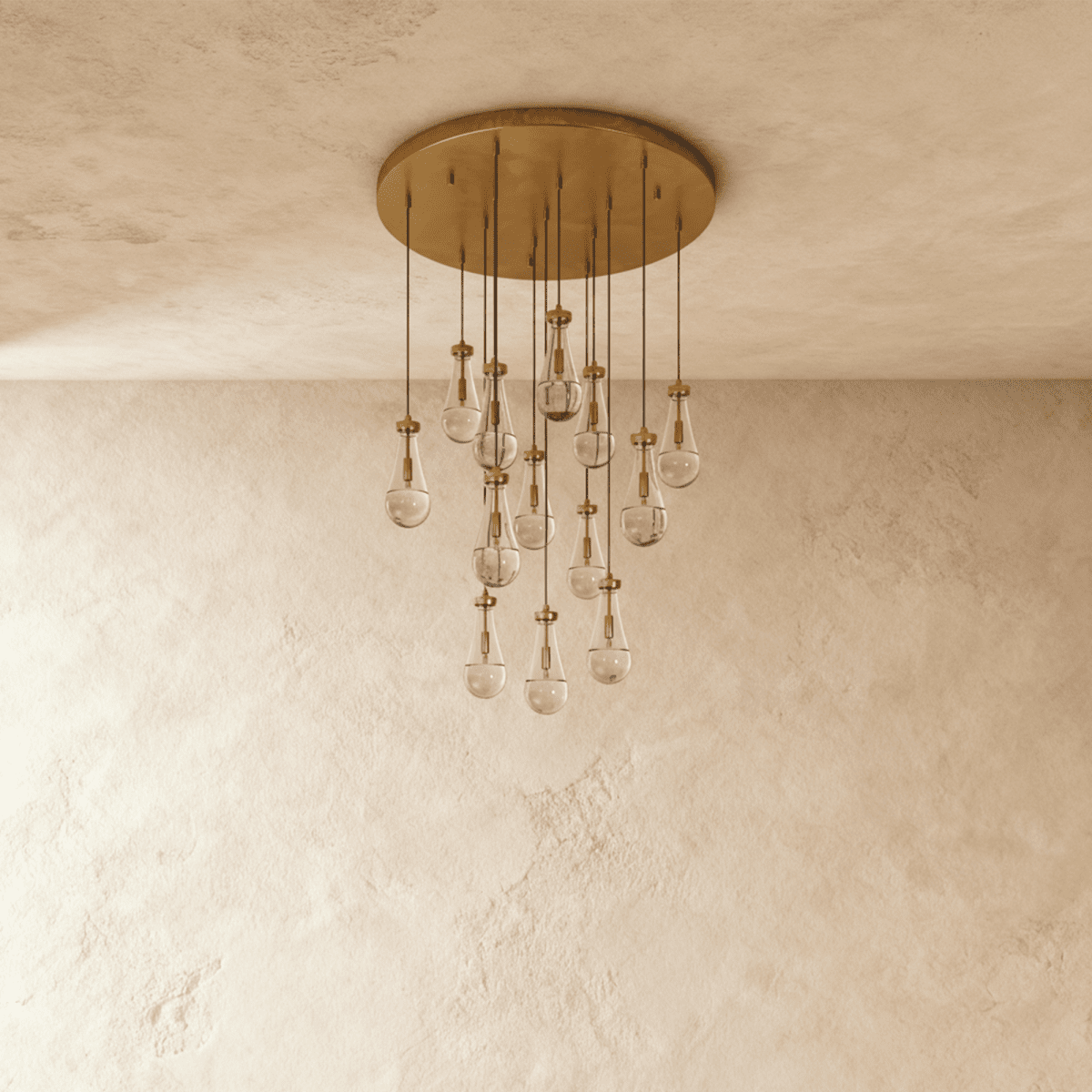 Clérachic chandelier with 13 clear glass teardrops on thin cords from a round brushed gold canopy, switched off, against limewashed plaster