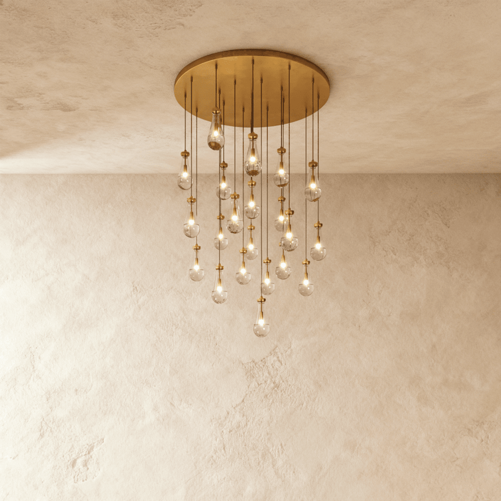 Clérachic chandelier with 21 clear glass teardrops on thin cords from a round brushed gold canopy, glowing warm, against limewashed plaster