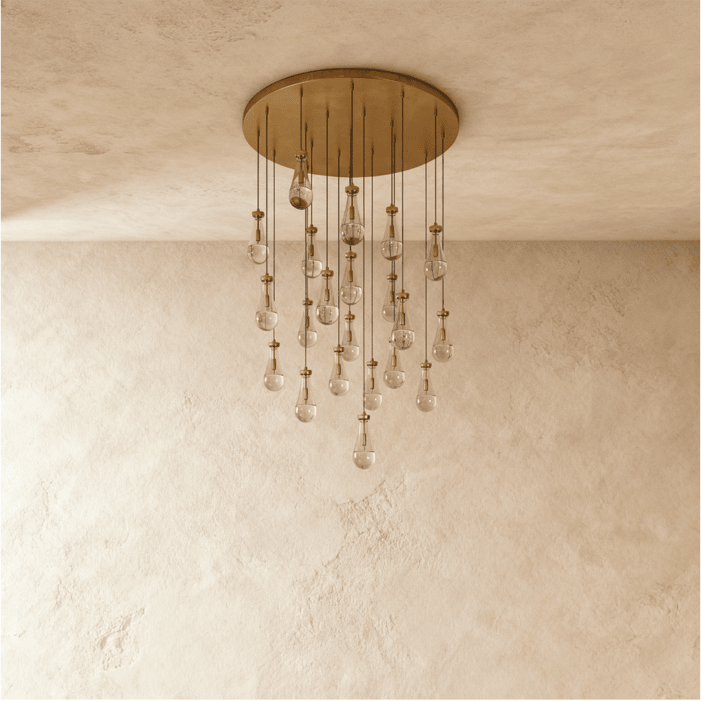Clérachic chandelier with 21 clear glass teardrops on thin cords from a round brushed gold canopy, switched off, against limewashed plaster