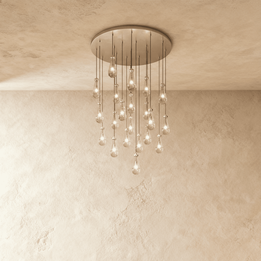 Clérachic chandelier with 21 clear glass teardrops on thin cords from a round satin nickel canopy, glowing warm, against limewashed plaster