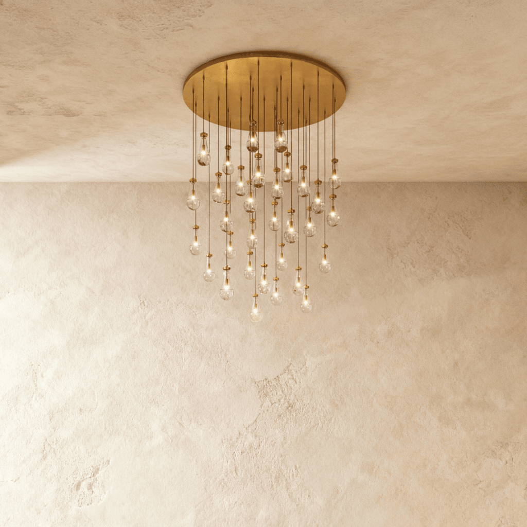 Clérachic chandelier with 31 clear glass teardrops on thin cords from a round brushed gold canopy, glowing warm, against limewashed plaster