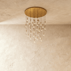 Clérachic chandelier with 31 clear glass teardrops on thin cords from a round brushed gold canopy, glowing warm, against limewashed plaster