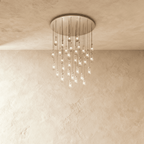 Clérachic chandelier with 31 clear glass teardrops on thin cords from a round satin nickel canopy, glowing warm, against limewashed plaster