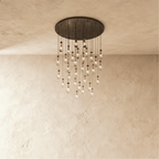 Clérachic chandelier with 31 clear glass teardrops on thin cords from a round dark bronze canopy, glowing warm, against limewashed plaster