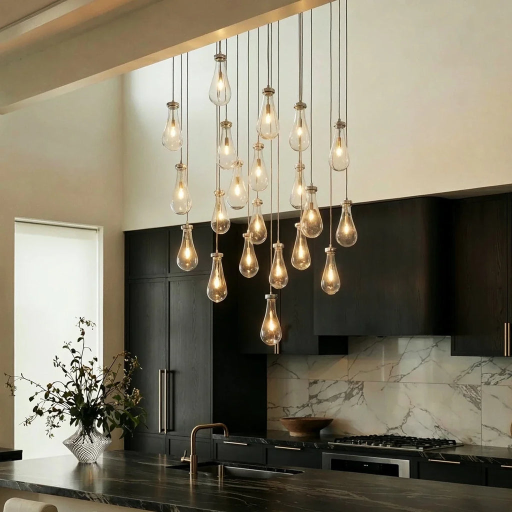 Clérachic chandelier with clear glass drops in a nickel finish, lit warm, above a kitchen island with dark cabinets and marble splashback