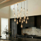 Clérachic chandelier with clear glass drops in a nickel finish, lit warm, above a kitchen island with dark cabinets and marble splashback