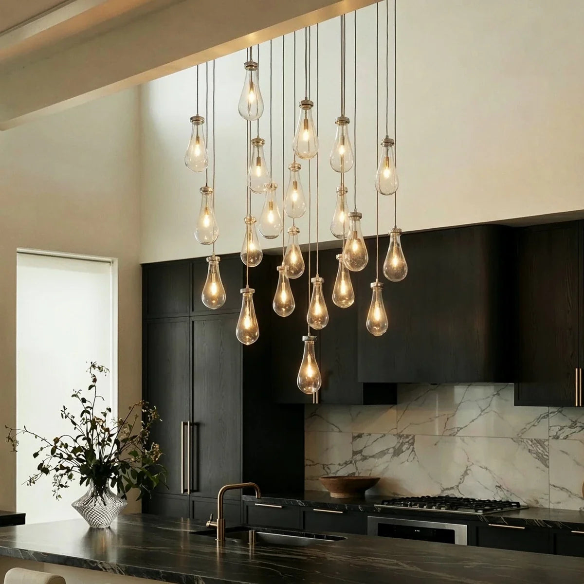 Clérachic chandelier with clear glass drops in a nickel finish, lit warm, above a kitchen island with dark cabinets and marble splashback