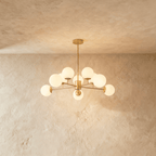 Clérachic gold chandelier with 9 milky white glass globes on straight arms, lit warm, against a limewash wall.