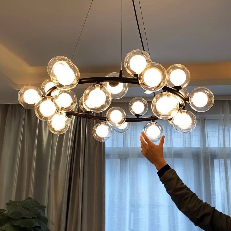 Clérachic black ring chandelier of clear glass globes with frosted inner bulbs, lit above a room with grey curtains and a raised hand.
