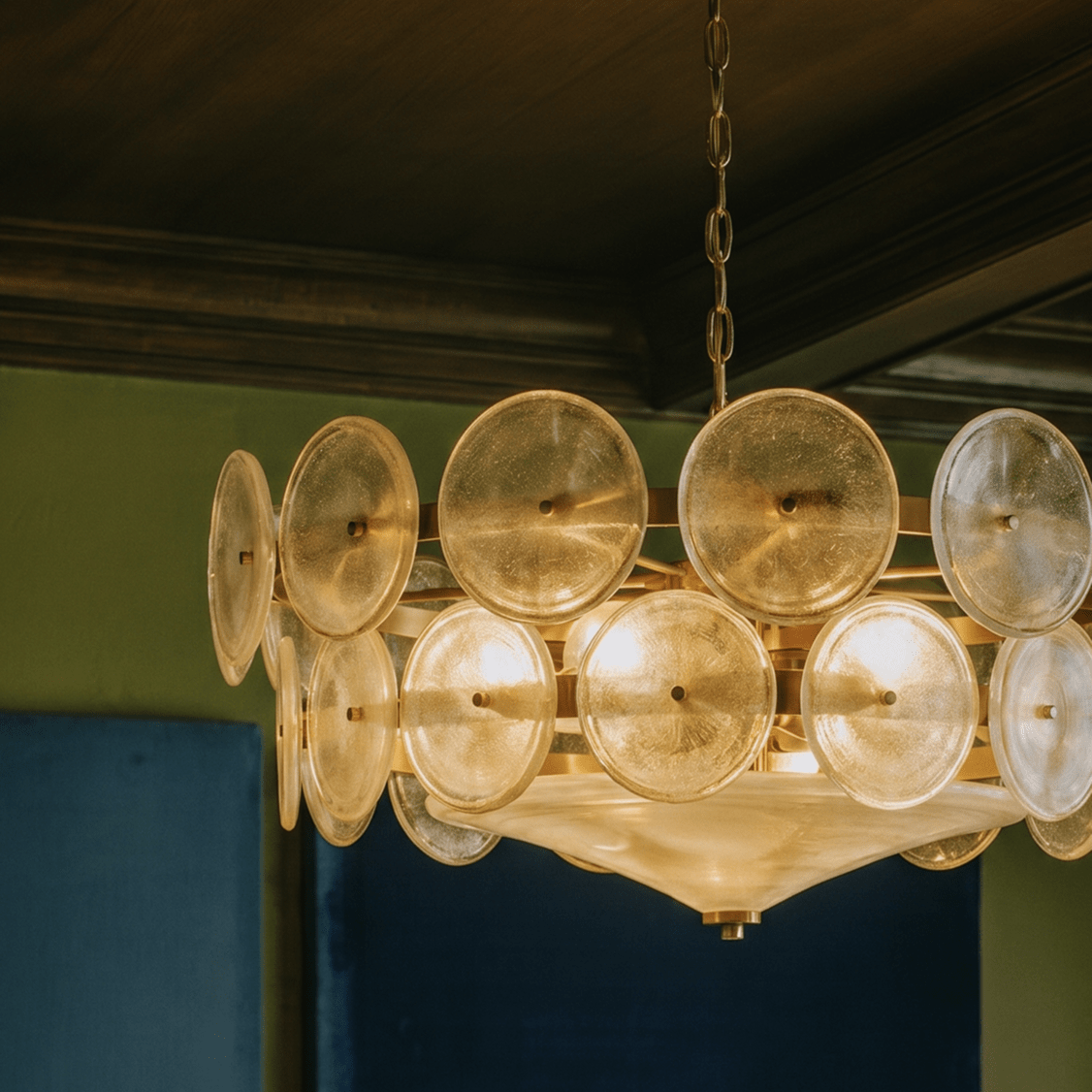 Clérachic chandelier glowing warm, its textured champagne glass discs and brass arms close up against a dark wood ceiling.