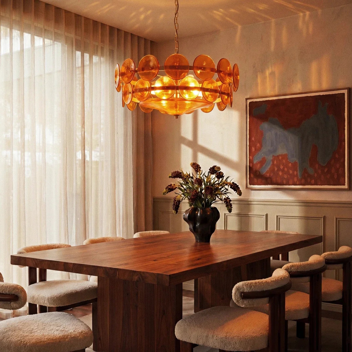 Clérachic chandelier with lit amber glass discs above a walnut dining table, with sheer curtains and wall art behind it.