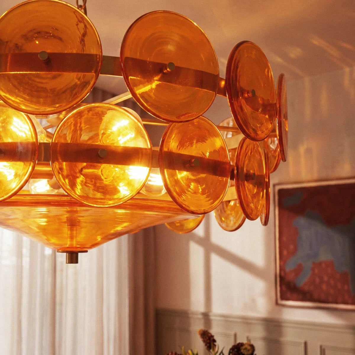 Clérachic chandelier close-up, glowing amber glass discs and bulbs inside the frame, with a dining room softly blurred behind.