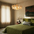 Clérachic disc chandelier lit warm above a bedroom with a green linen bed, linen curtains, a bedside table and landscape art.