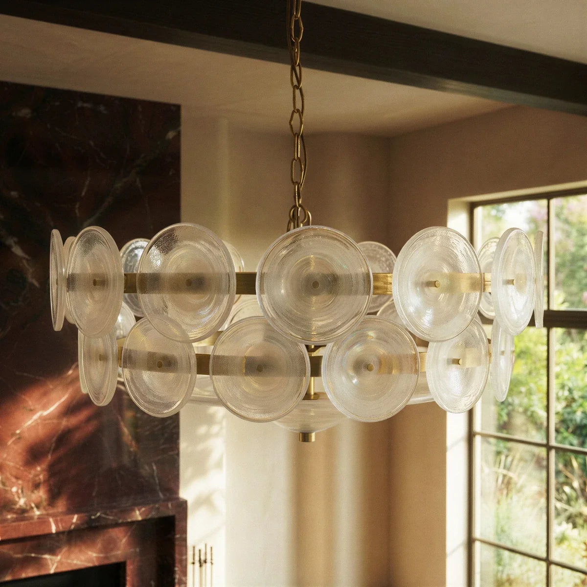 Clérachic chandelier close-up of clear ribbed glass discs on brass bands, in a room with a dark marble fireplace and window.
