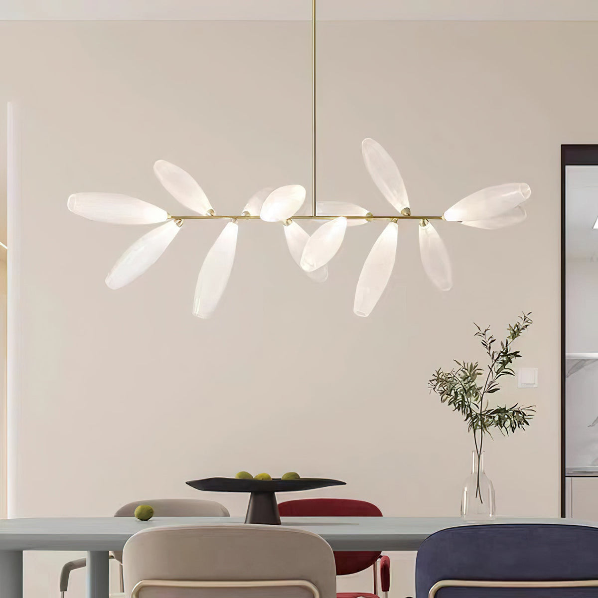 A long Clérachic chandelier with frosted white glass leaves on a gold rod hangs above a dining table with pale chairs in a calm cream room.