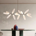 A linear Clérachic chandelier of white glass leaves on a gold bar hangs over a dark green marble table with a bowl and sculptural vases.