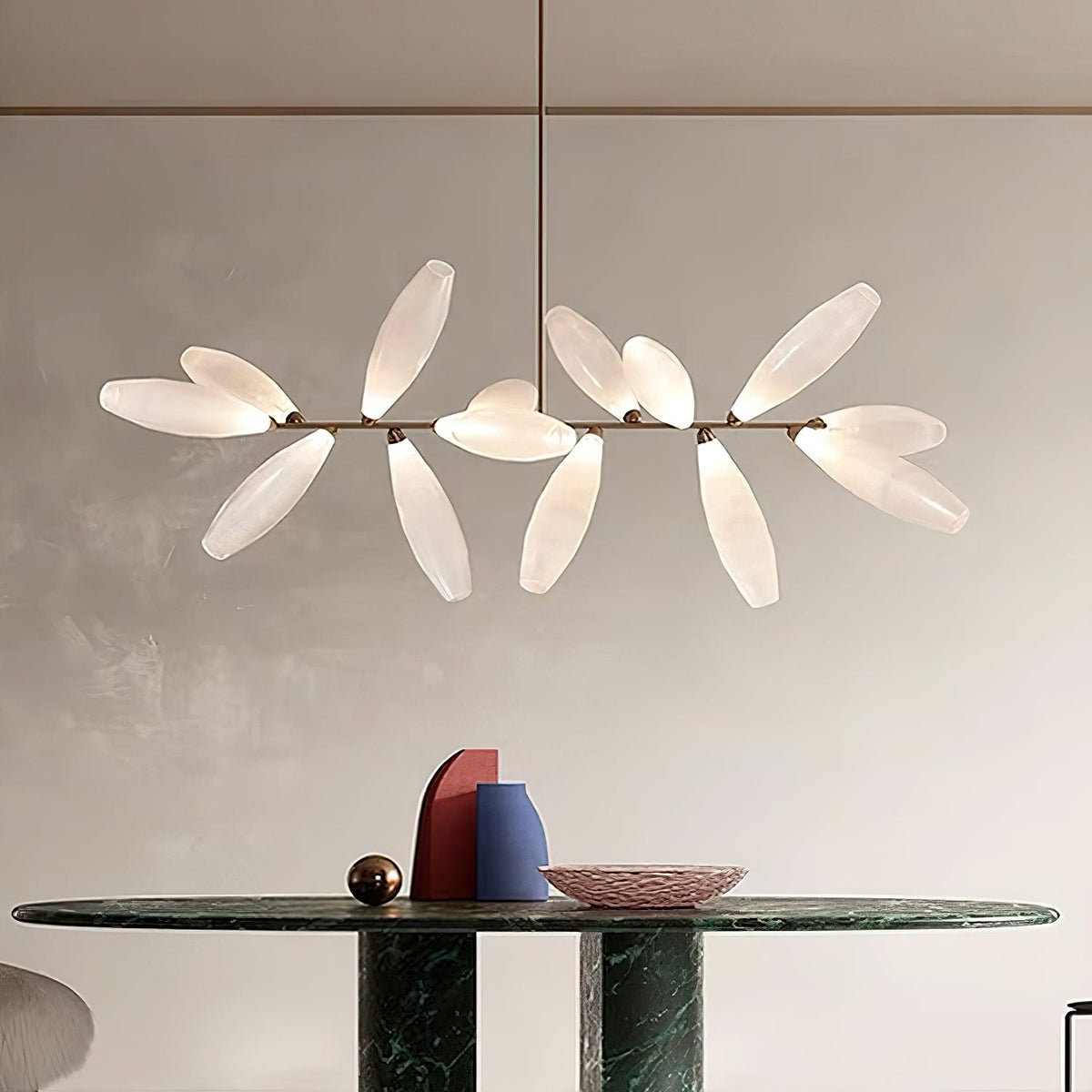 A linear Clérachic chandelier of white glass leaves on a gold bar hangs over a dark green marble table with a bowl and sculptural vases.