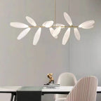 A linear Clérachic chandelier of white glass leaves hangs above a table with a dark runner, a gold ornament and a pink pleated dining chair.