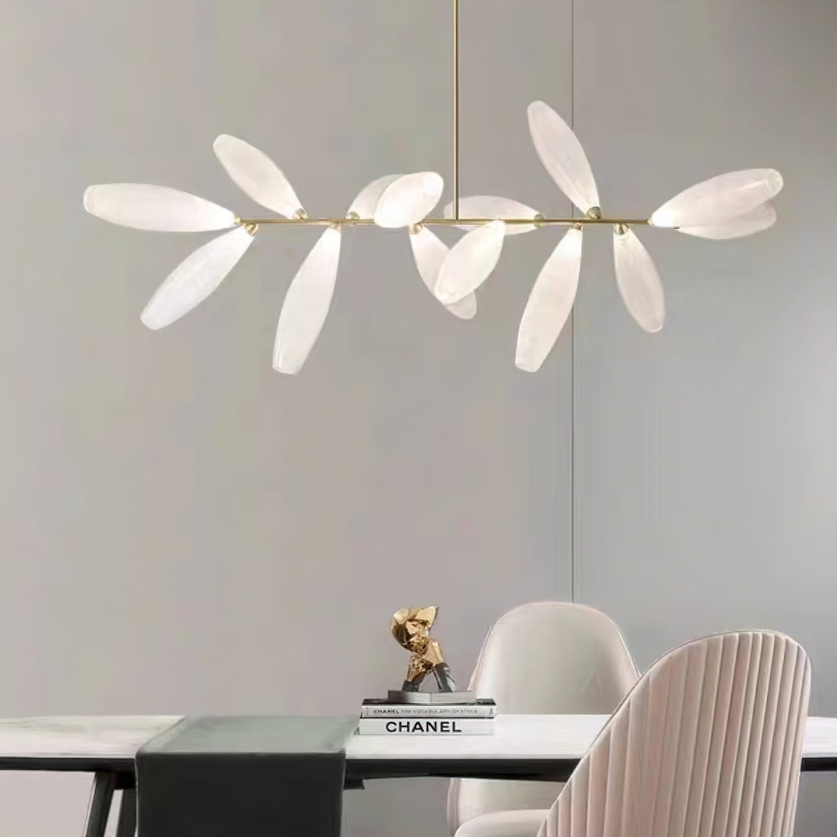A linear Clérachic chandelier of white glass leaves hangs above a table with a dark runner, a gold ornament and a pink pleated dining chair.
