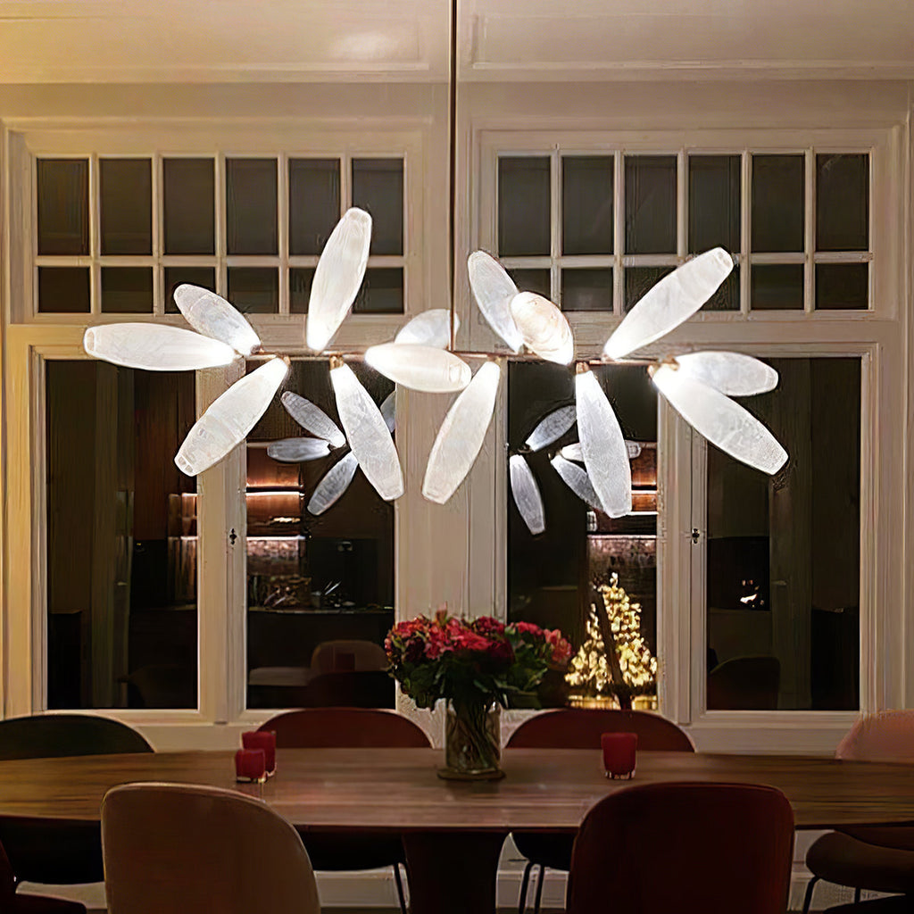 A Clérachic chandelier with lit white glass leaves hangs over a round dark wood table with red flowers, in front of tall glazed doors at night.