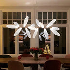 A Clérachic chandelier with lit white glass leaves hangs over a round dark wood table with red flowers, in front of tall glazed doors at night.