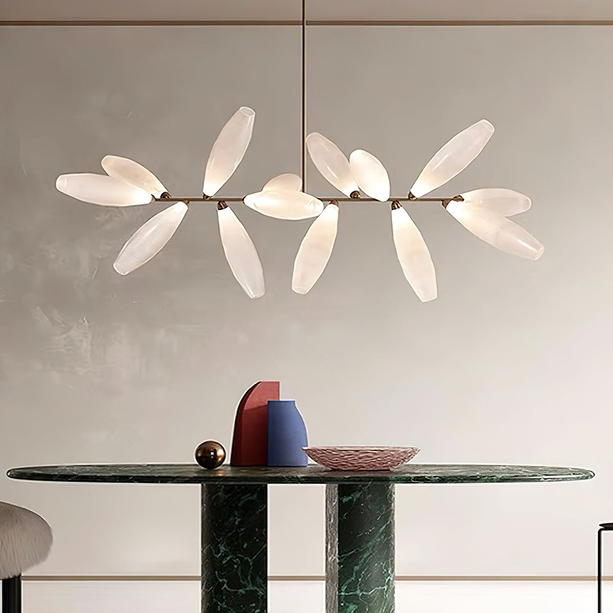 A linear Clérachic chandelier of frosted glass leaves on a gold bar hangs above a dark green marble table and a pink dish.