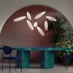 Several Clérachic glass leaves hang at different heights on slim gold stems in an arched, plum-coloured room with a teal table and large plants.