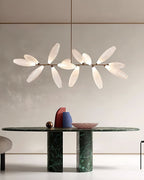 A linear Clérachic chandelier of frosted glass leaves on a gold bar hangs above a long dark green marble table in a pale room.