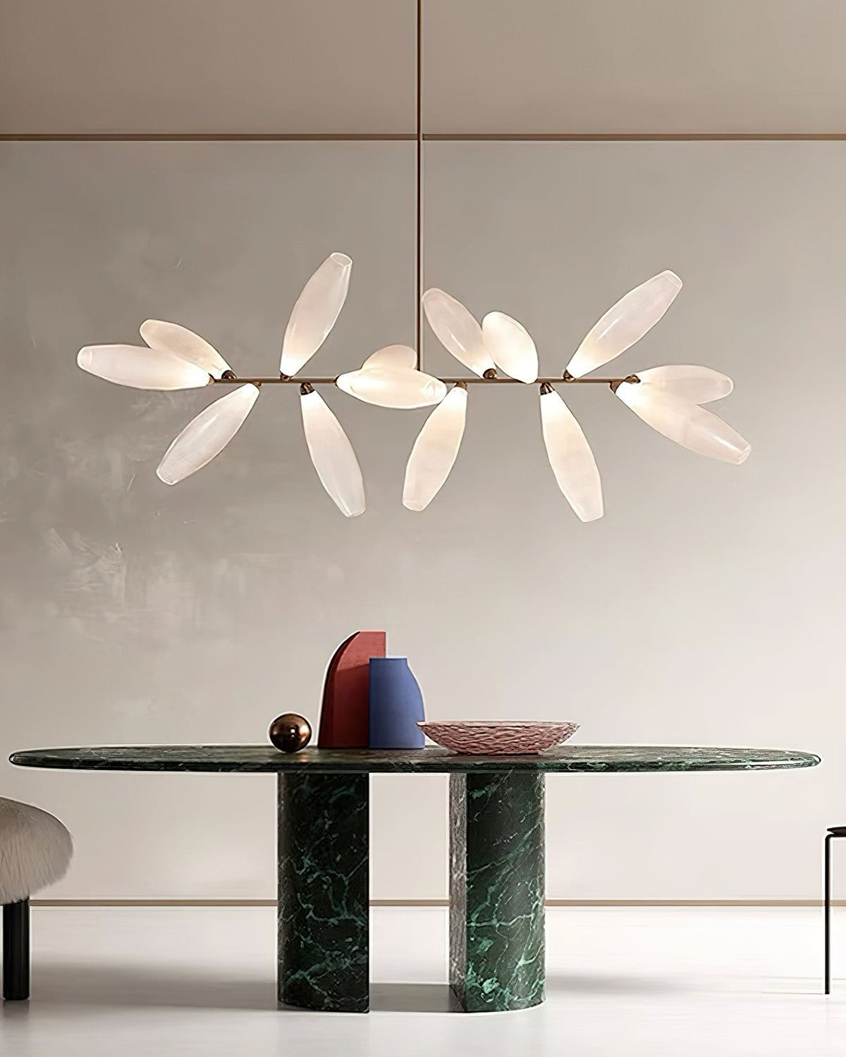 A linear Clérachic chandelier of frosted glass leaves on a gold bar hangs above a long dark green marble table in a pale room.