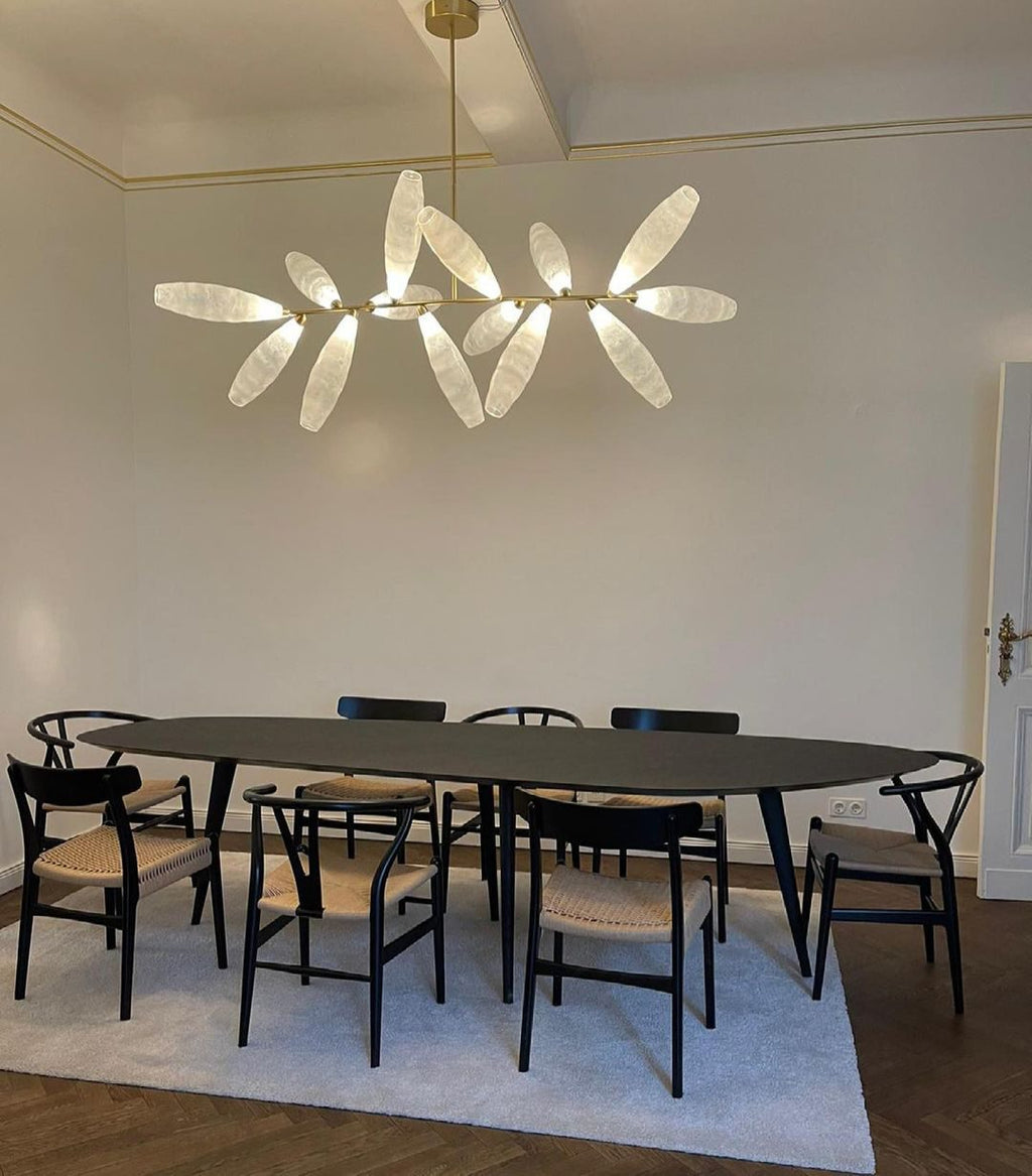 A Clérachic chandelier of white glass leaves hangs over a long black oval table with woven-seat wooden chairs and a pale rug.